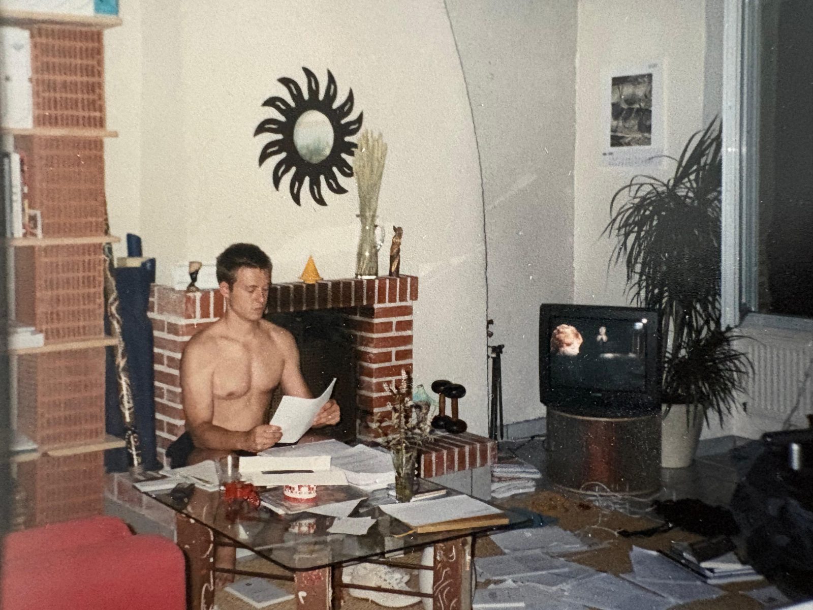 A print from the thesis years: Nicolas Boitout reading a sheet of paper at a glass table in front of a brick fireplace, with books, notes and loose pages spread over the table and across the floor around him.